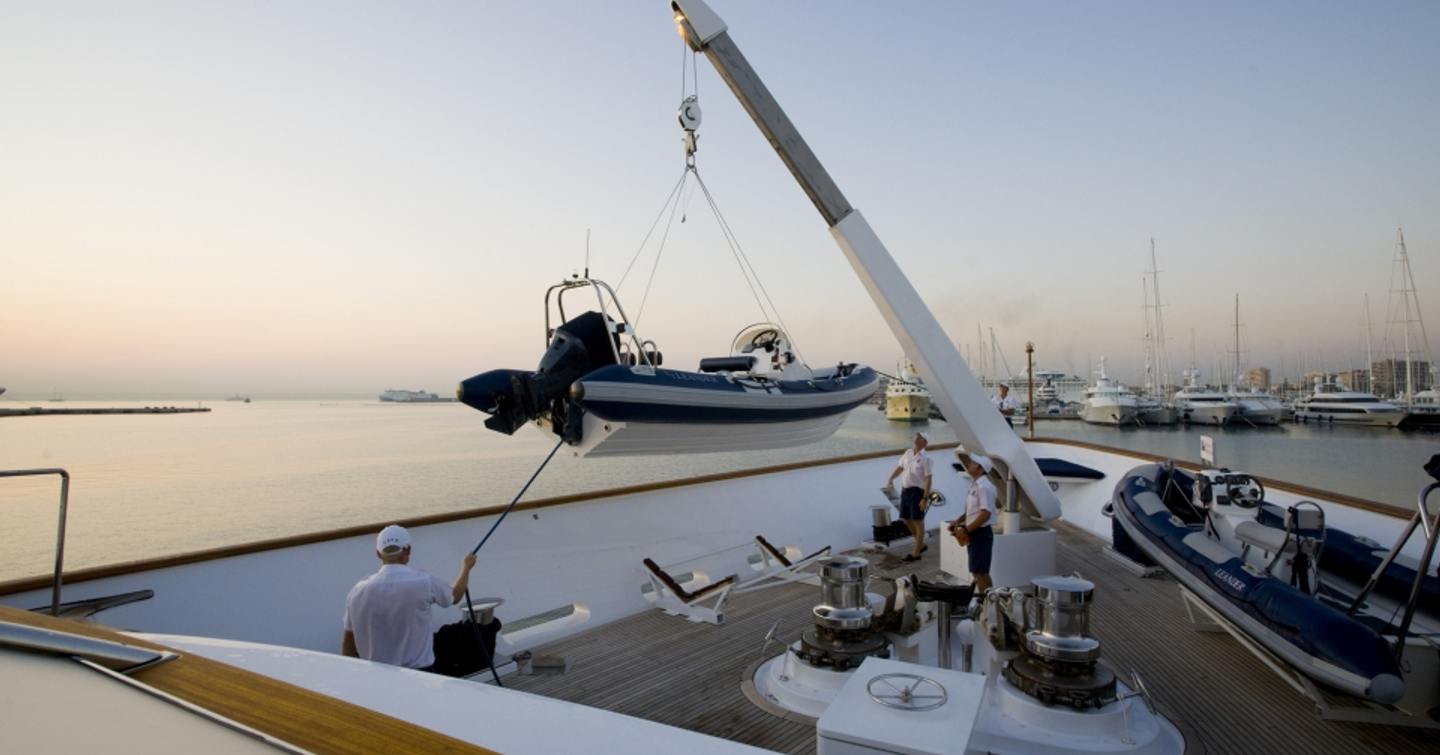 RIB tender being lifted by crane onto main deck of yacht at sunset with three crew members helping