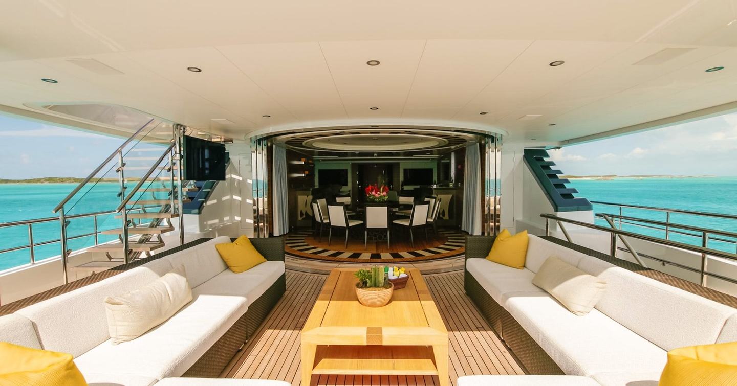 Yacht aft deck with two white sofas, yellow cushions, wooden coffee table, and dining area overlooking turquoise water.