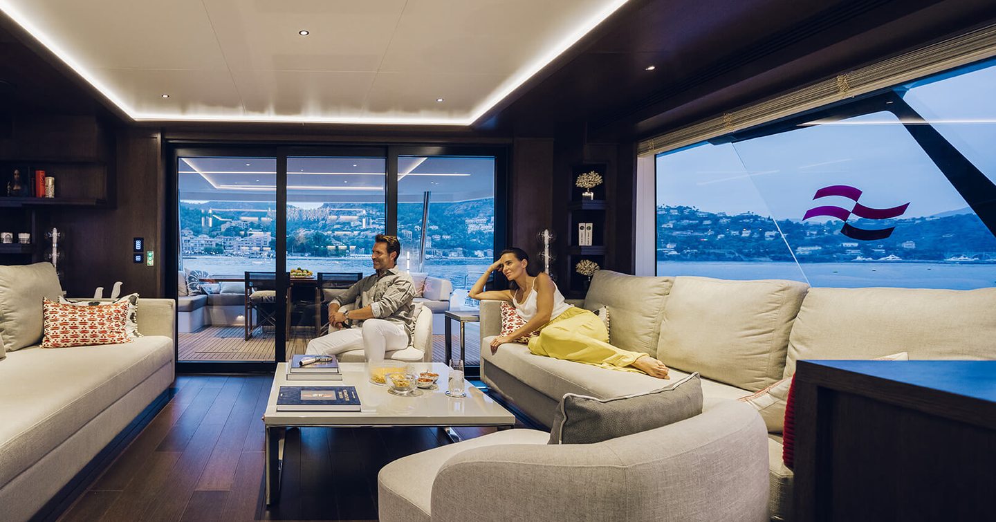 Main salon onboard Sirena Yachts 88, facing white sofas and coffee table with man and lady relaxing and watching TV