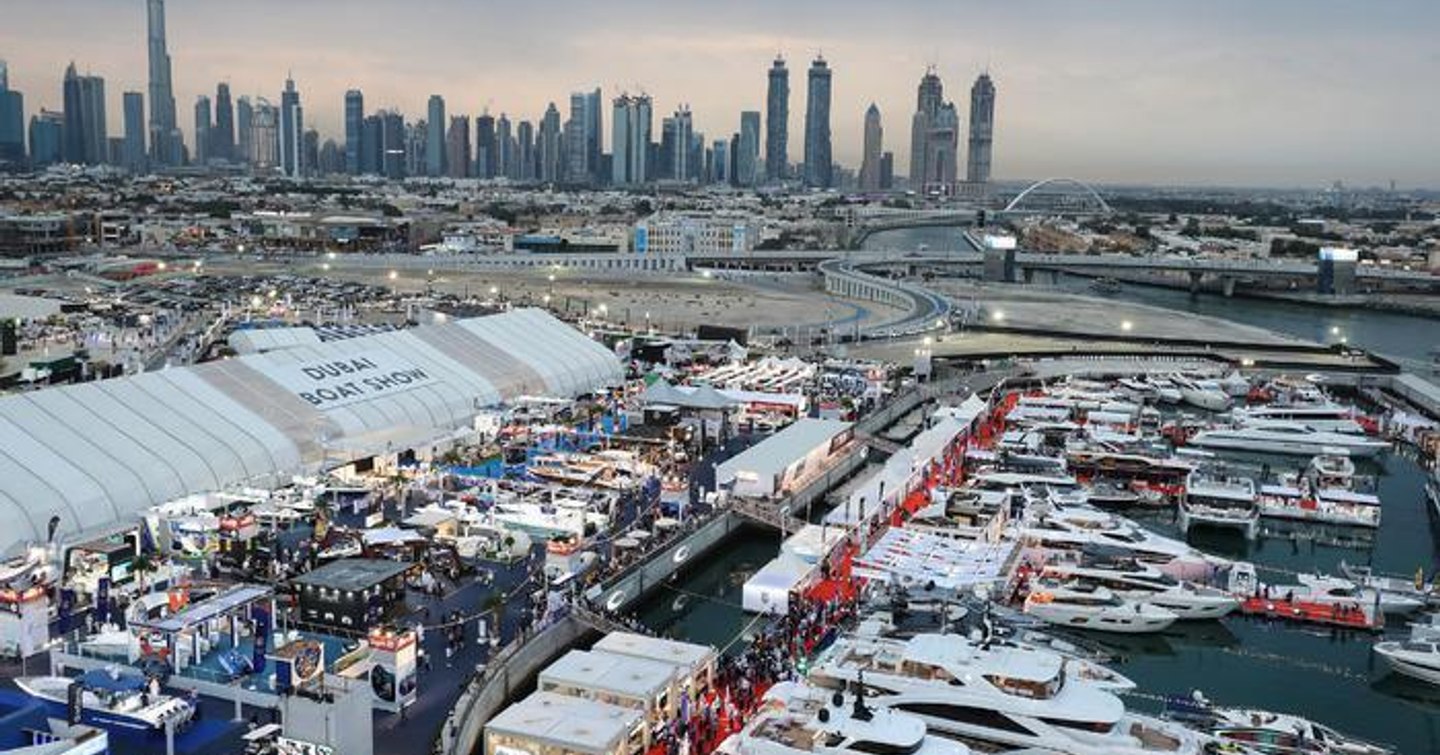 Overview of Dubai International Boat Show, multiple yachts moored along docks with Dubai skyscrapers in background.