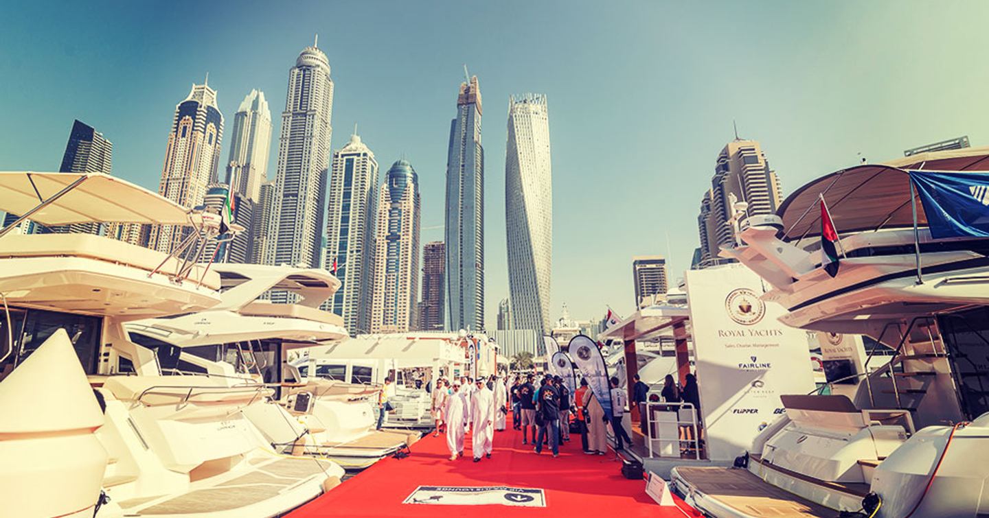 Overview at ground level of Dubai boat show, looking up towards skyscrapers, with yachts berthed either side of red carpeted slip.