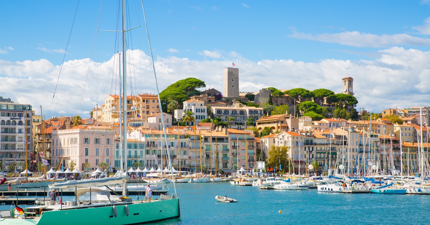 Overview of Cannes Vieux Port, sailing boats and motor yachts berthed in marina, overlooking Cannes in background.