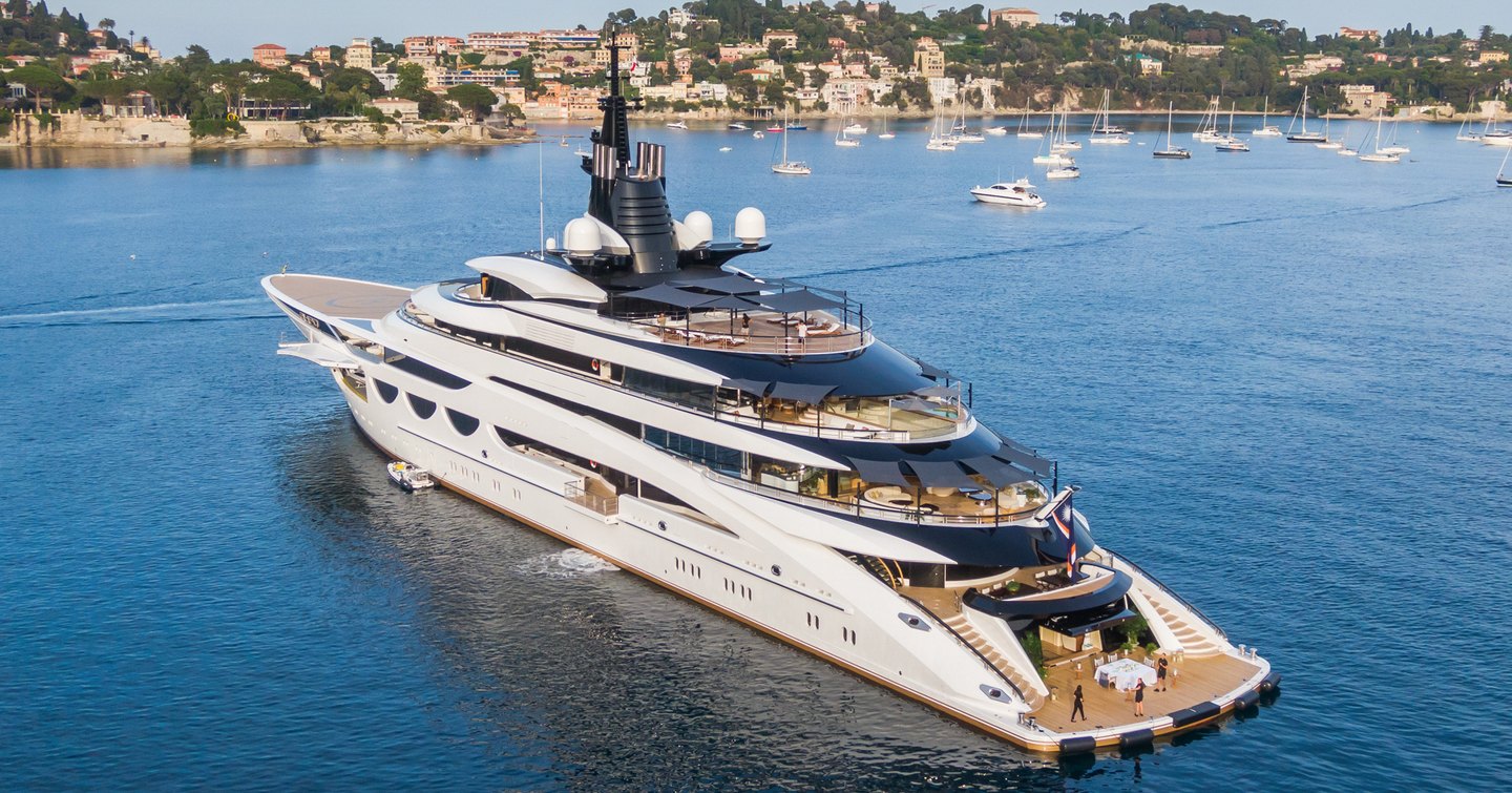 Aft view of superyacht Lady Jorgia, all exterior decks visible with the sea all around the yacht, plus a Mediterranean settlement in the background.