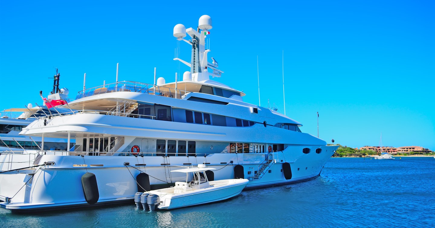 Tender boat in front of large yacht that is docked on clear water with clear blue sky in background