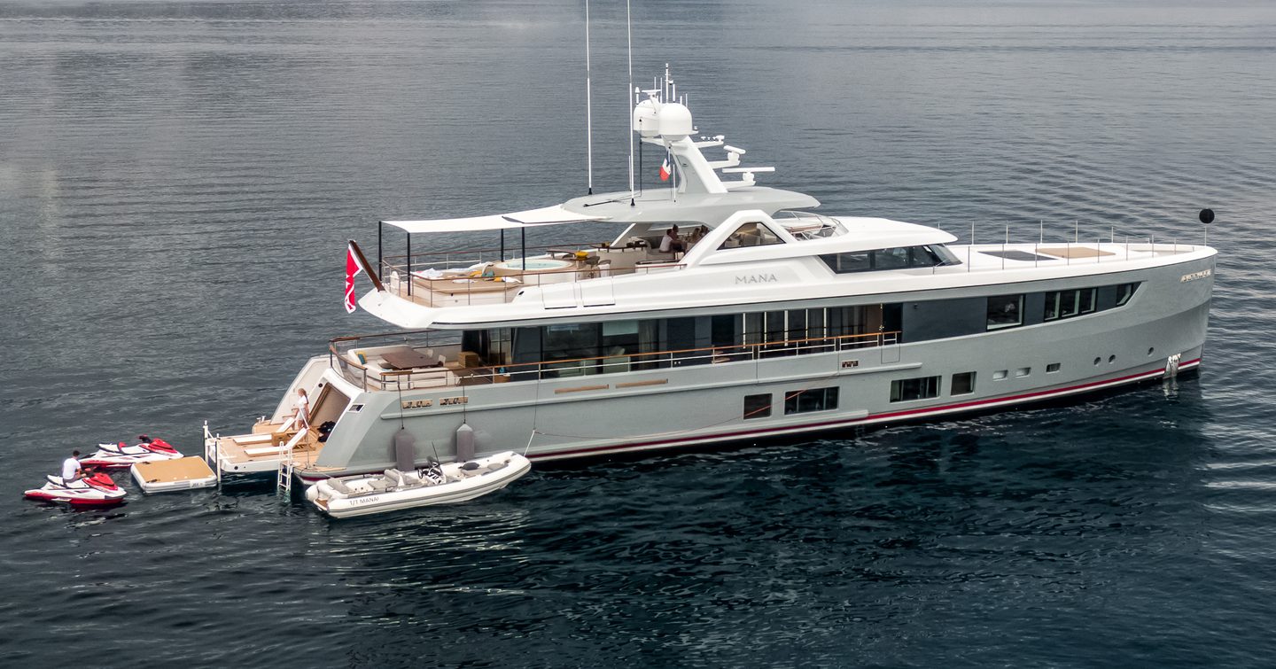 Mulder ThirtySix MANA I shown mid aerial over starboard quarter and side profile, the yacht is on anchor in still waters with land in background. a tender is berthed alongside the yacht and the bathing platform is extended with personal watercraft in the water