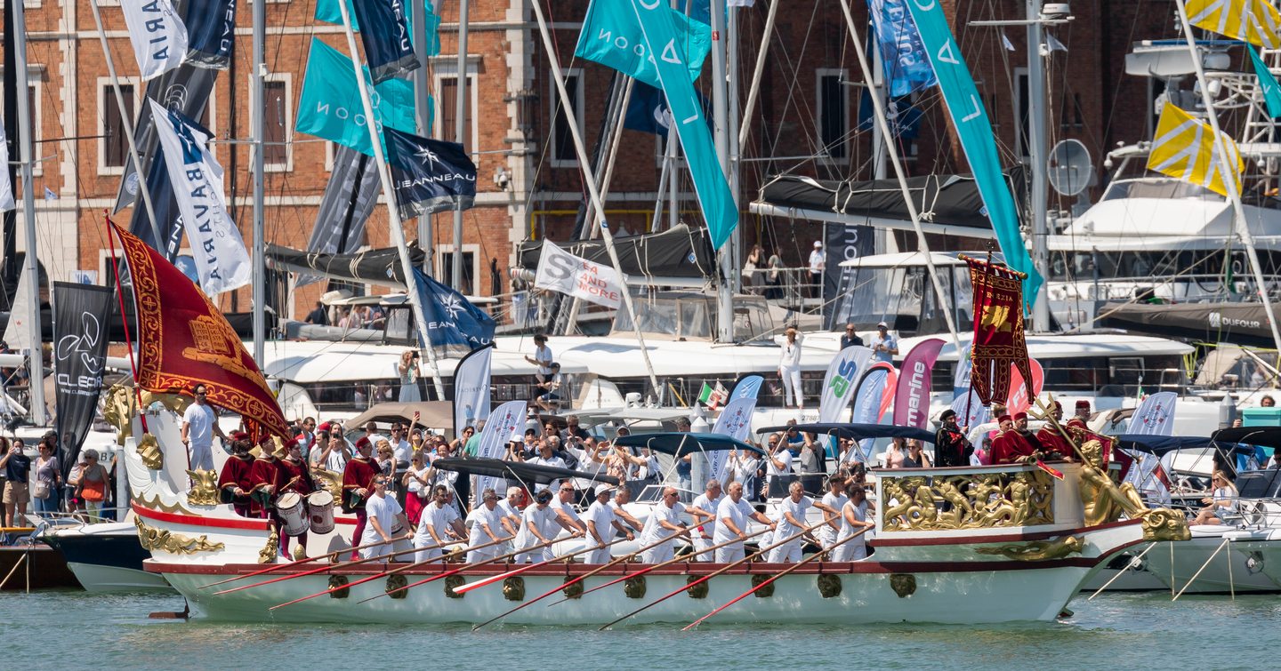 A regatta in action at the Venice Boat Show, with a wooden boat being rowed in the foreground and flags behind