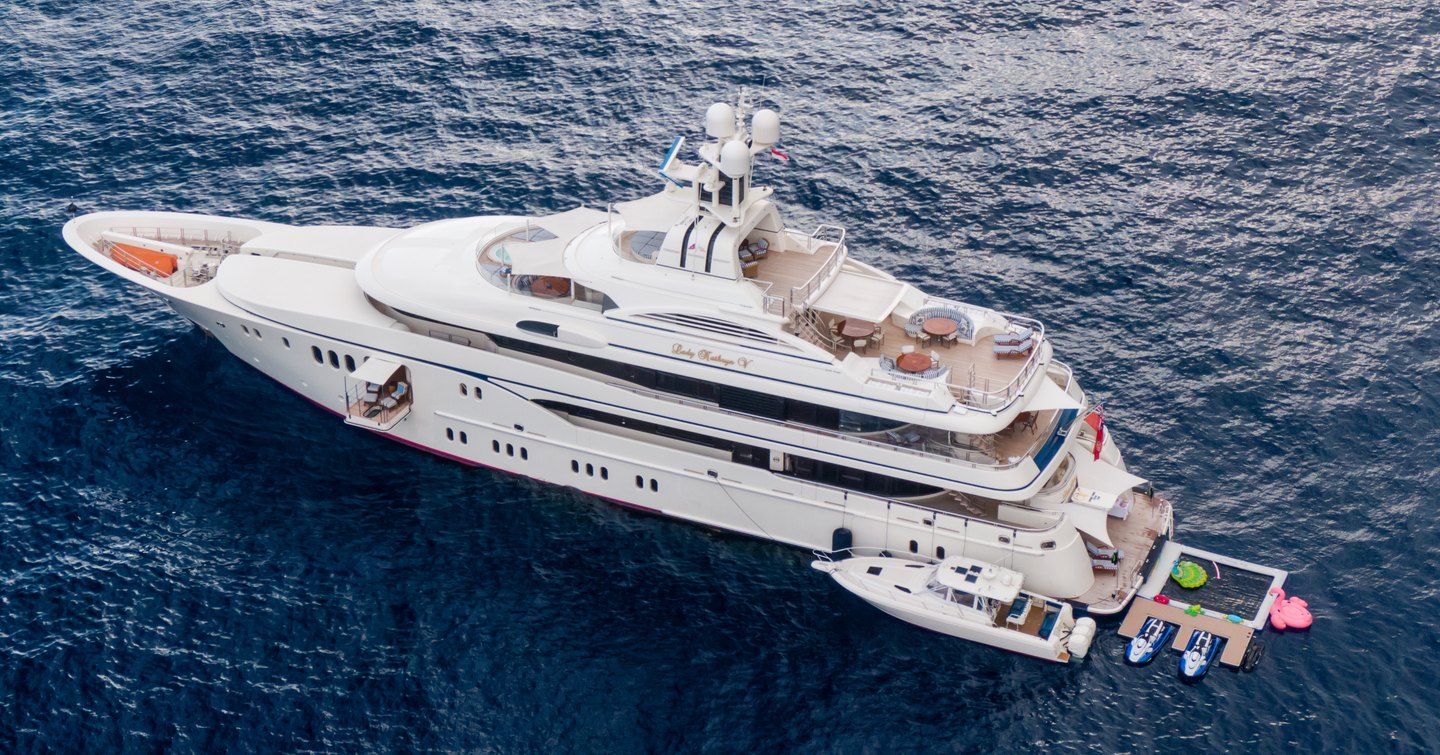 Lurssen yacht LADY KATHRYN V shown in aerial short over port side, the yacht is stationary as it has the tender moored alongside and the aft deck bathing platform appears busy with toys