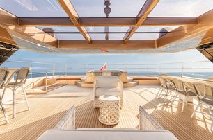 100 Sunreef Power exterior deck shot taken from inside, looking out over freestanding furniture over a mirror like sea, on a very bright day, the light coming through the roof panels casting shadows on the smart teak deck