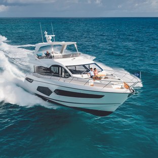 Manhattan 68 running shot on clear day, mid aerial view over starboard bow with couple relaxing on sun pads, person on flybridge helm