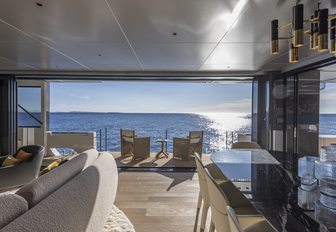 Arcadia A96 motor yacht interior salon looking out through large open glazing doors to the exterior seating area, sun low in sky casting reflection over water