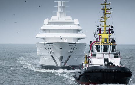 Forward view of Amels 120 being escorted by a tug boat in the foreground, surrounded by sea.