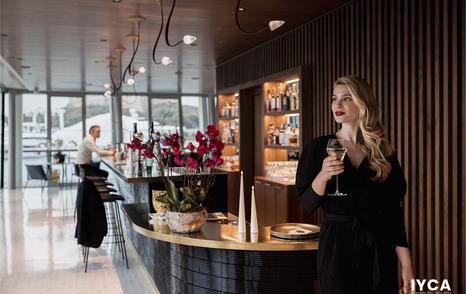 Bar at the International Yacht Club d’Antibes (IYCA). Man sitting on a stool at the bar, with woman in foreground holding glass of wine.