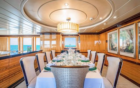 Motor yacht Nita K II's elegant yacht dining room with long table set for eight, cream upholstered chairs, large chandelier, and windows overlooking the ocean