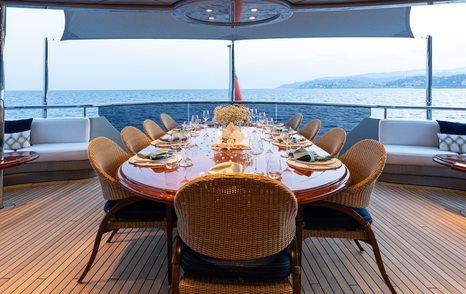 Motor yacht Vibrance's aft deck dining area with set table, wicker chairs, and sea view at sunset.
