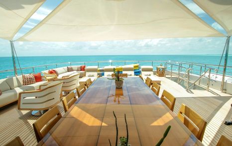 Motor yacht W's aft deck with large dining table, lounge seating, and canopy shade overlooking turquoise ocean views.