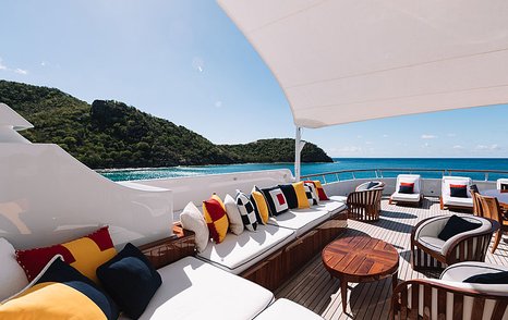 Exterior lounge area onboard Feadship superyacht FLAG, white seating with vibrant scatter cushions, surrounded by sea and elevated terrain in background.