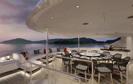 Exterior dining area onboard Heesen superyacht ATINA. Circular table and chairs adjacent to long sofas, boasting views over water and distant elevated terrain.