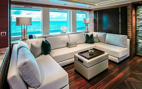 Motor yacht W's main saloon with white sectional sofa, dark accent pillows, wooden floors, and large windows overlooking the ocean.
