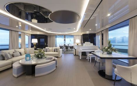 Main salon onboard Mangusta Oceano 50. Monochrome design with plush seating to port and grand piano visible to starboard. Surrounded by large windows.