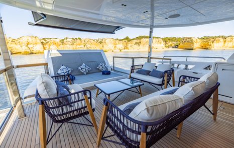 Exterior dining area onboard Sunseeker 100 Yacht, wicker sofas and armchairs facing in towards a coffee table on outside deck, surrounded by sea with cliffs in background.