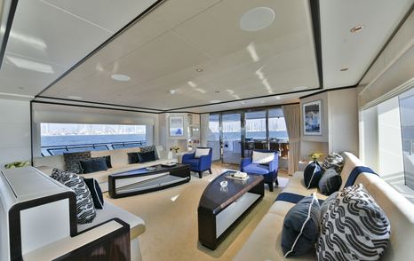 Main salon onboard the Majesty 120. White angular seating around the exterior walls with curved coffee tables centre. Surrounded by wide windows.