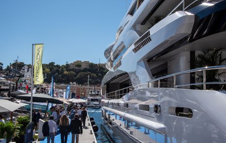 Aft view of a superyacht berthed at the Monaco Yacht Show with visitors walking along pontoon to port.