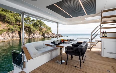 Aft deck onboard Sirena 78, alfresco dining table with sofa one side and dining chairs the other. Stairs to flybridge visible.