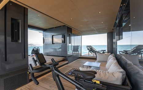 Overview of the beach club onboard superyacht PANAM. Gym equipment and sofa visible inside with sun lounger sat on fold down balcony in background.