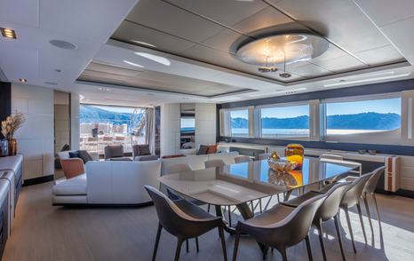 Interior dining area onboard Pershing 140 'Touch Me' , glass table with 10 chairs in foreground, surrounded by wide windows and a lounge area aft.