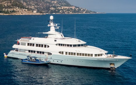 Feadship yacht OLYMPUS on anchor in calm sea on bright day, yacht tender berthed alongside starboard side, land in background