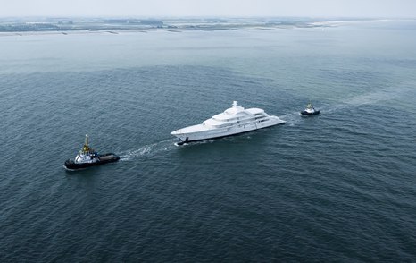 Amels 120 being escorted by tug boats on route to Vlissingen, surrounded by water with land in far distance.