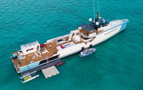 Damen Yacht Support 5009 SHADOW aerial view over starboard quarter deck and side profile, stationary in calm waters, all shell doors appear to be open, with tenders and watercraft moored alongside