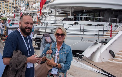 Visitors to the Monaco Yacht Show smiling at photographer with large superyachts moored in background.