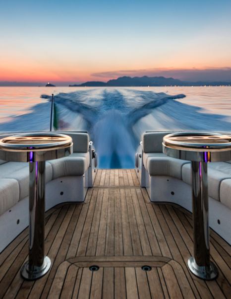 Teak decking and two tables with cream sofas on either side on aft of Wooden Boats WB14 limousine tender. Sunset and sea in the background.