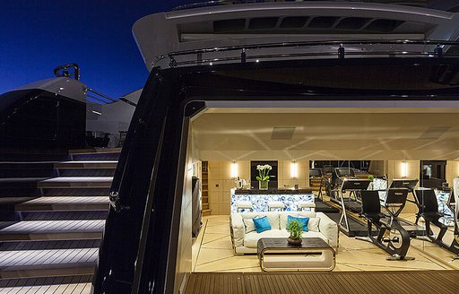 Overview of the beach club onboard M/Y JULIA. Gym equipment and sofa visible in open transom area, with steps leading up to main deck on portside.