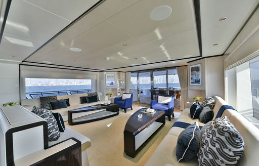 Main salon onboard the Majesty 120. White angular seating around the exterior walls with curved coffee tables centre. Surrounded by wide windows.