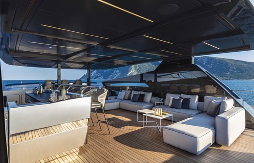 Overview of shaded flybridge onboard Pershing 140 'Touch Me', U shaped sofa arrangement facing foreground with wetbar and stools to port side, surrounded by sea and cliffs in distance