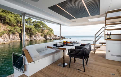 Aft deck onboard Sirena 78, alfresco dining table with sofa one side and dining chairs the other. Stairs to flybridge visible.