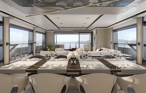 Overview of interior dining area onboard Majesty 111. Long table with white chairs horizantal in foreground with lounge area visible behind.