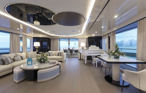 Main salon onboard Mangusta Oceano 50. Monochrome design with plush seating to port and grand piano visible to starboard. Surrounded by large windows.