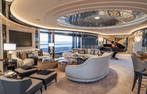 Overview of interiors onboard motor yacht KISMET, with plush cream seating and a grand piano in the background