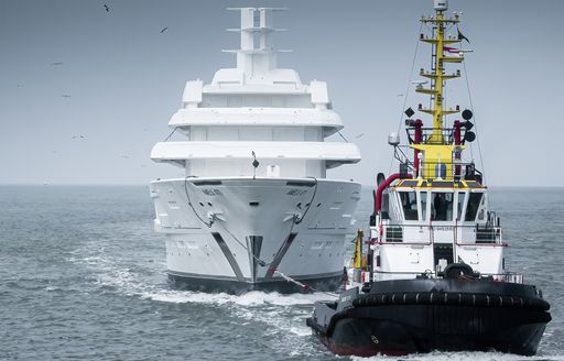 Forward view of Amels 120 being escorted by a tug boat in the foreground, surrounded by sea.