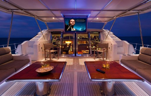 Two tables undercover on yacht deck with large TV screen visible above glass doors.