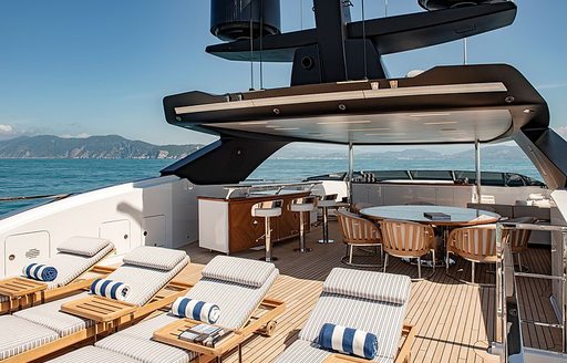 Sun deck onboard Sanlorenzo Lady Lena, four sun loungers in foreground, overlooking a wetbar to port side and round dining table starboard, surrounded by sea