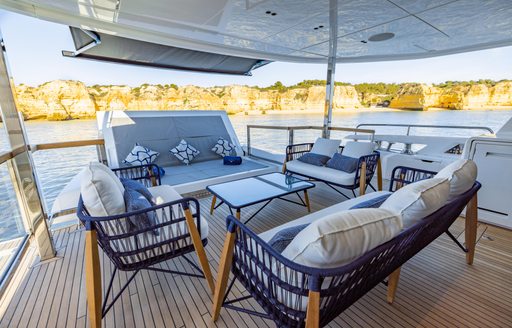 Exterior dining area onboard Sunseeker 100 Yacht, wicker sofas and armchairs facing in towards a coffee table on outside deck, surrounded by sea with cliffs in background.