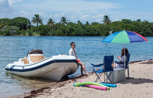 Man standing against RIB tender on shore of beach talking to female sitting on chair under umbrella