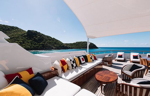 Exterior lounge area onboard Feadship superyacht FLAG, white seating with vibrant scatter cushions, surrounded by sea and elevated terrain in background.