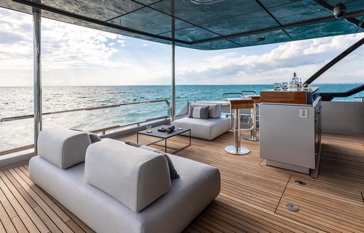 bar with bar stools and sofas on the teak-lined aft deck of the Argo 90 with cloudy blue sky beyond