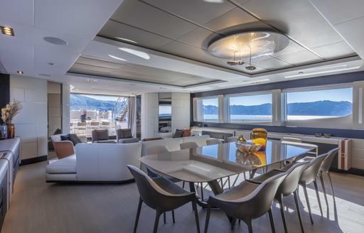 Interior dining area onboard Pershing 140 'Touch Me' , glass table with 10 chairs in foreground, surrounded by wide windows and a lounge area aft.