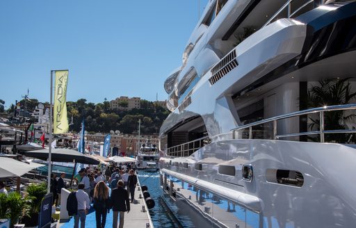 Aft view of a superyacht berthed at the Monaco Yacht Show with visitors walking along pontoon to port.